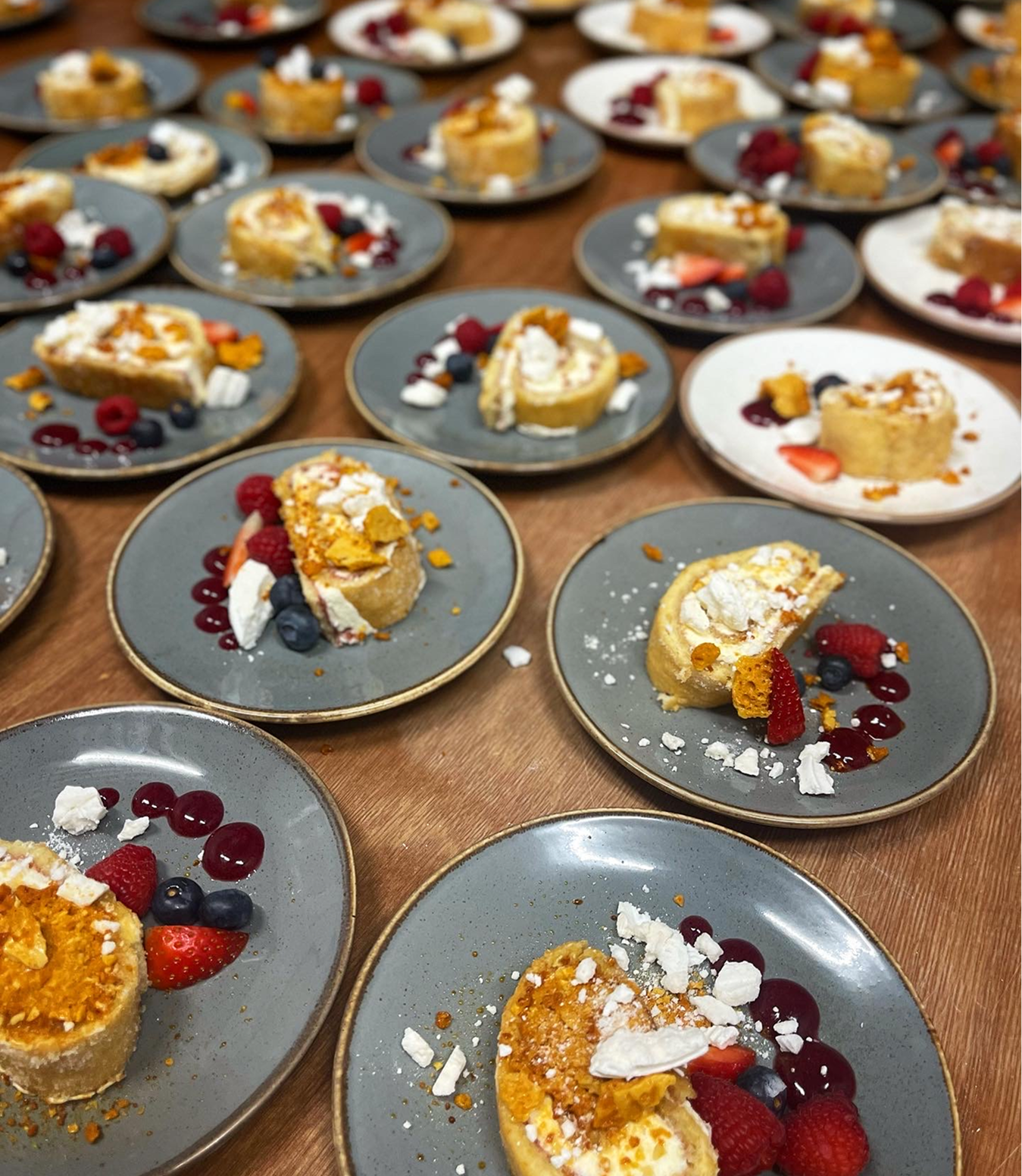 Multiple small plates with dessert, including roulades filled with cream and topped with berries and crumbled cookies on a wooden table.