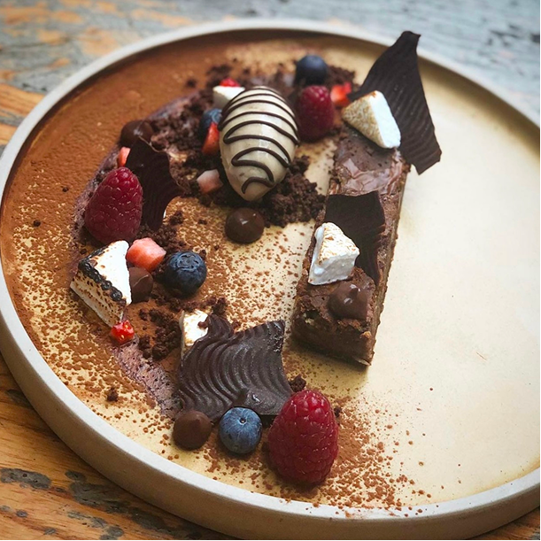 Chocolate tart garnished with raspberries, blueberries, small candies, and various pieces of chocolate, served on a round plate.