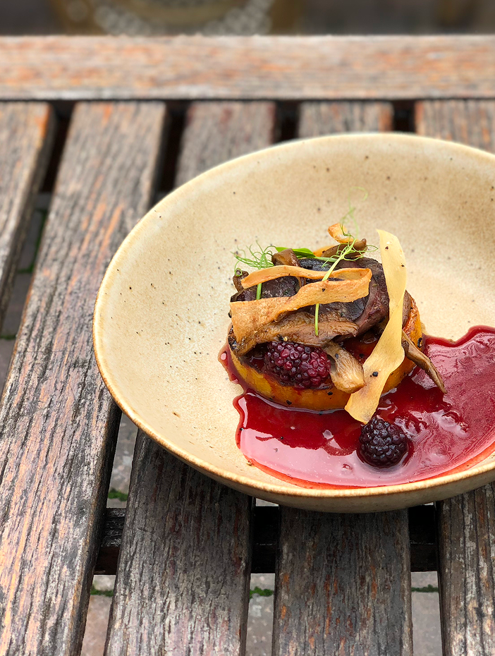 A ceramic bowl containing a gourmet dish with roasted vegetables, berries, and a red sauce, placed on a weathered wooden table outdoors.