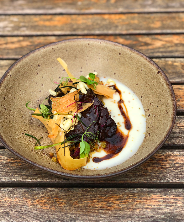 A gourmet dish in a beige bowl on a wooden surface, featuring vegetables, microgreens, and sauces.