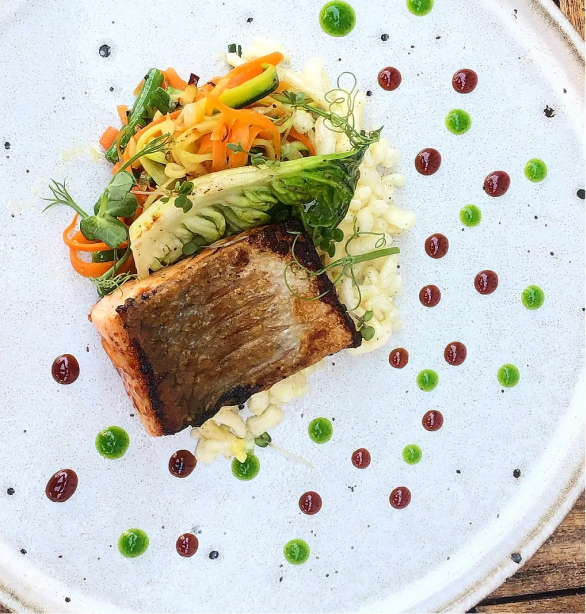 A plated dish with a piece of grilled fish, mashed potatoes, and sautéed mixed vegetables topped with microgreens, garnished with red and green sauce dots.