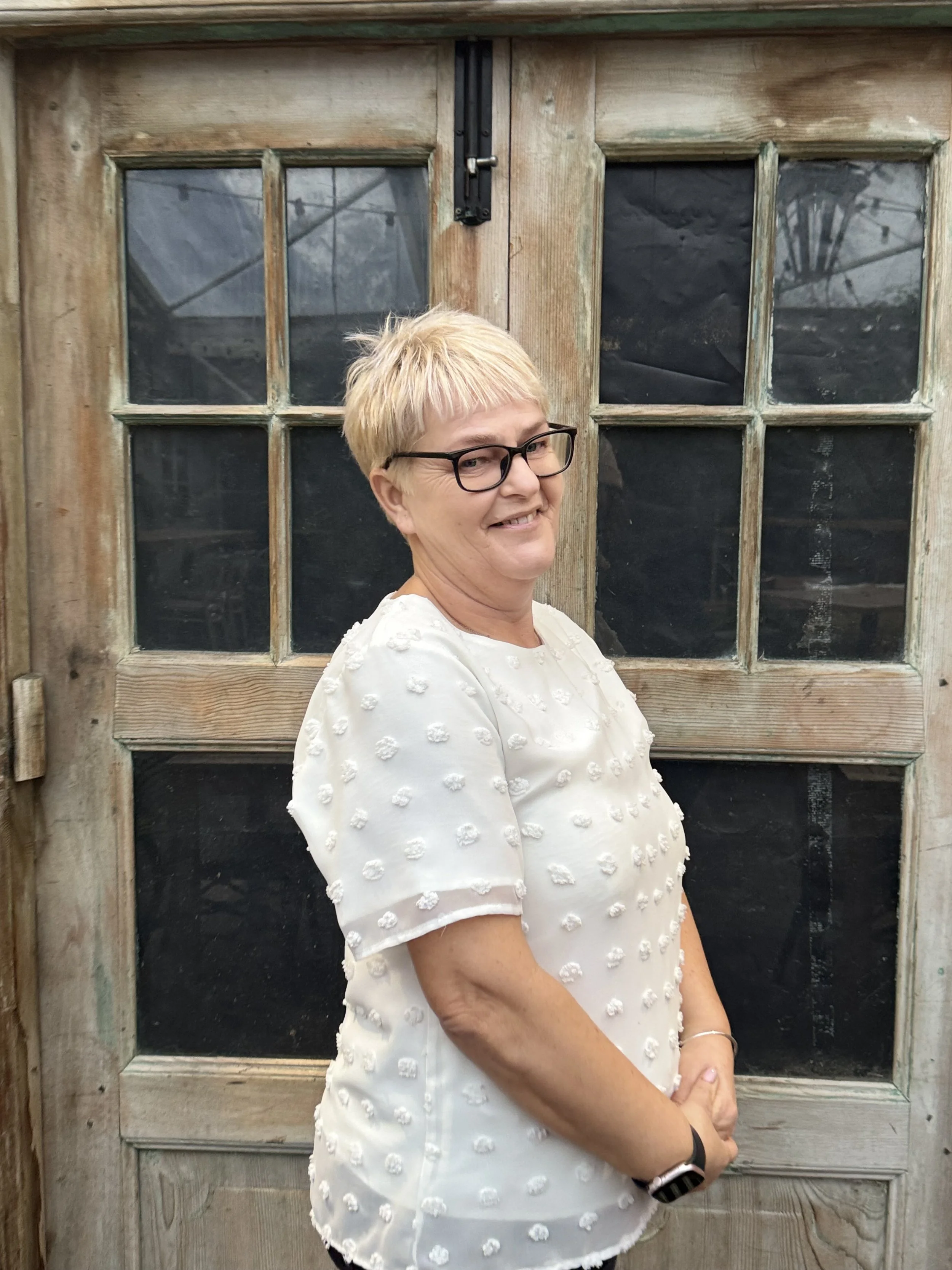 A woman with shoulder-length wavy brown hair, wearing a beige sleeveless top and beige pants, sitting on a bench with a white cushion, writing in a notebook. She is next to a wooden wall, with abstract artwork leaning against the wall nearby, in a room illuminated by sunlight.