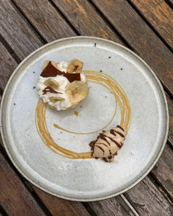 Dessert plate with whipped cream topped with banana slices, chocolate pieces, and nuts, drizzled with caramel and chocolate sauces, on a wooden table.