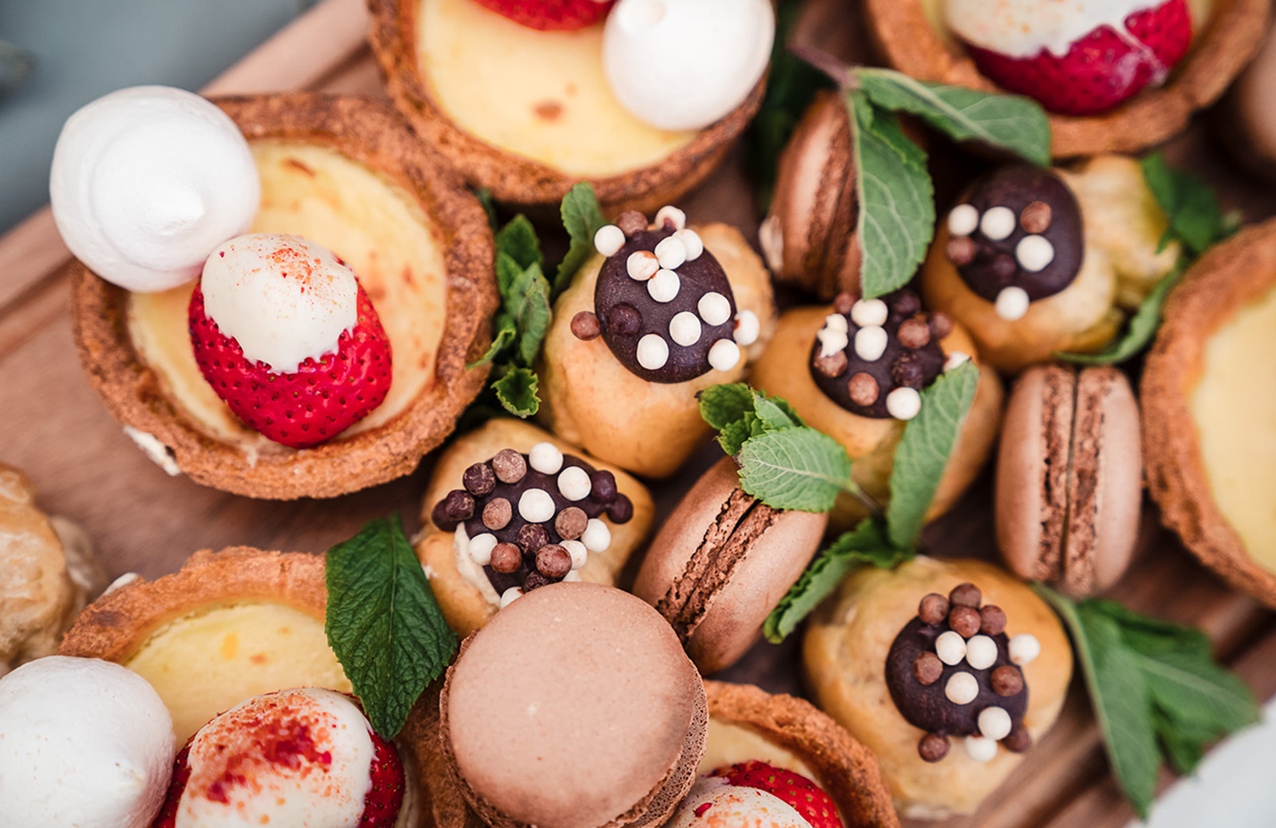 Assorted mini desserts including tart shells with strawberries and cream, macarons, decorated chocolates with sprinkles, and garnished with fresh mint leaves.
