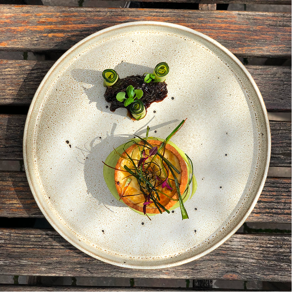 A plate with a gourmet appetizer, featuring a round slice of melon topped with thin green vegetable strips and microgreens, and a small pile of dark tapenade garnished with green onion curls.