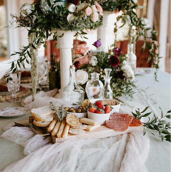 An elegant table setting with a cheese platter, strawberries, and olives, surrounded by floral arrangements and glass decanters, on a white tablecloth.