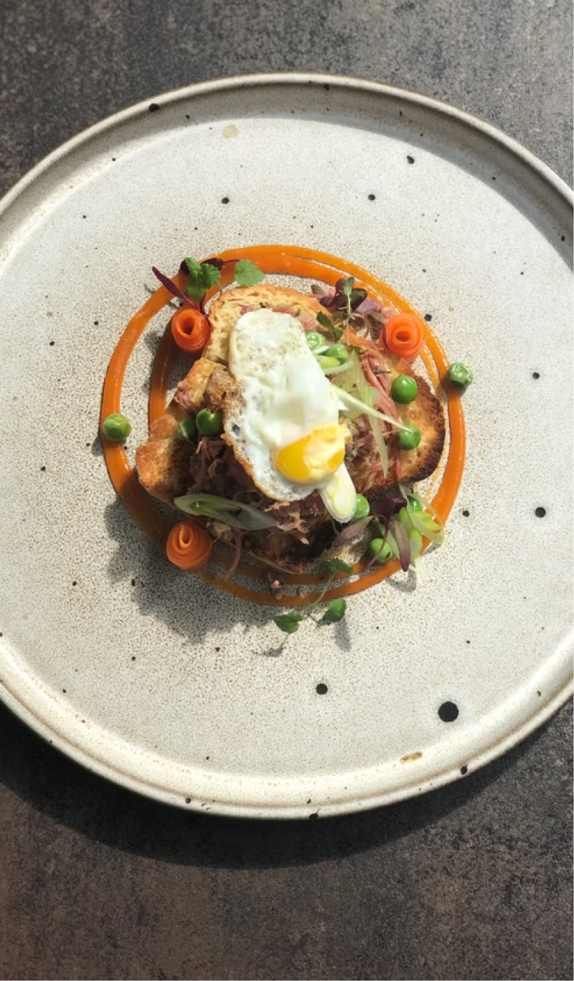A plate with a gourmet dish featuring a slice of toasted bread topped with shredded meat, a poached egg, and garnished with microgreens, with a decorative swirl of orange sauce and small dollops of green sauce.
