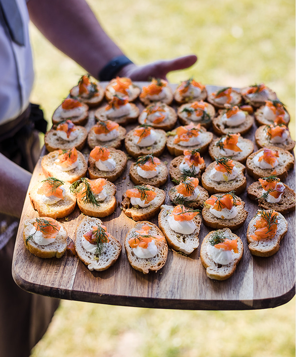 A person holding a wooden tray filled with small bread slices topped with cream, smoked salmon, and garnished with herbs, likely at an outdoor event.