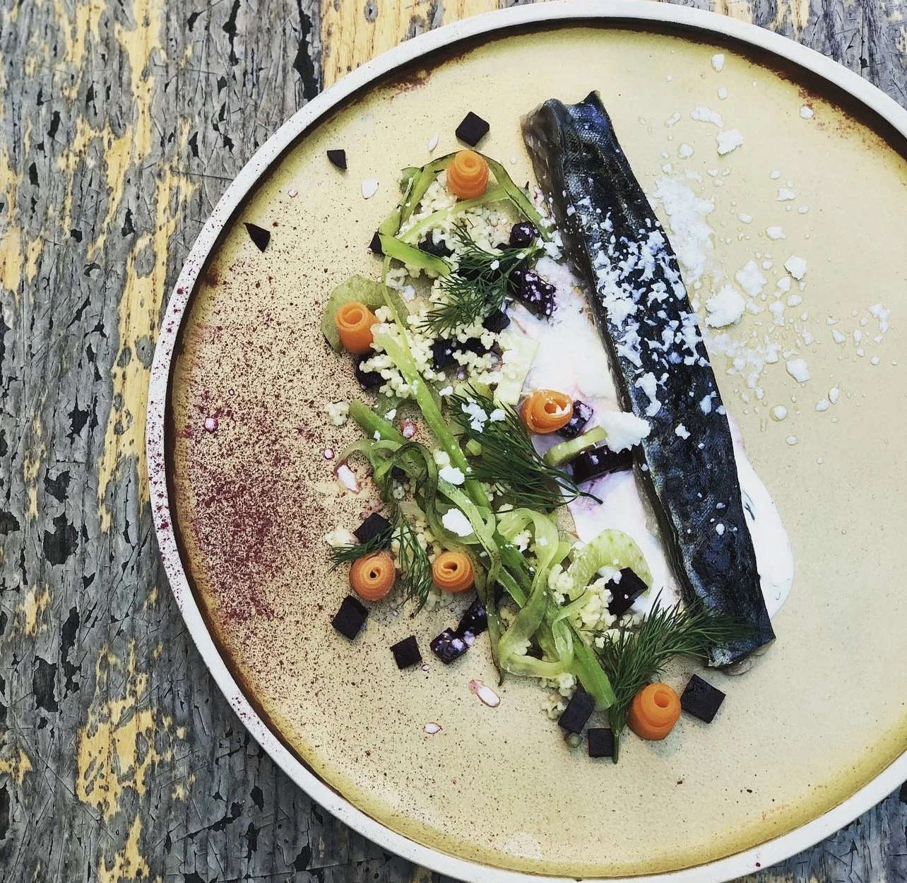 A plated dish featuring a strip of blackened fish on a bed of sauce, garnished with thinly sliced vegetables, herbs, small diced vegetables, and black and orange decorative elements, served on a speckled beige plate.