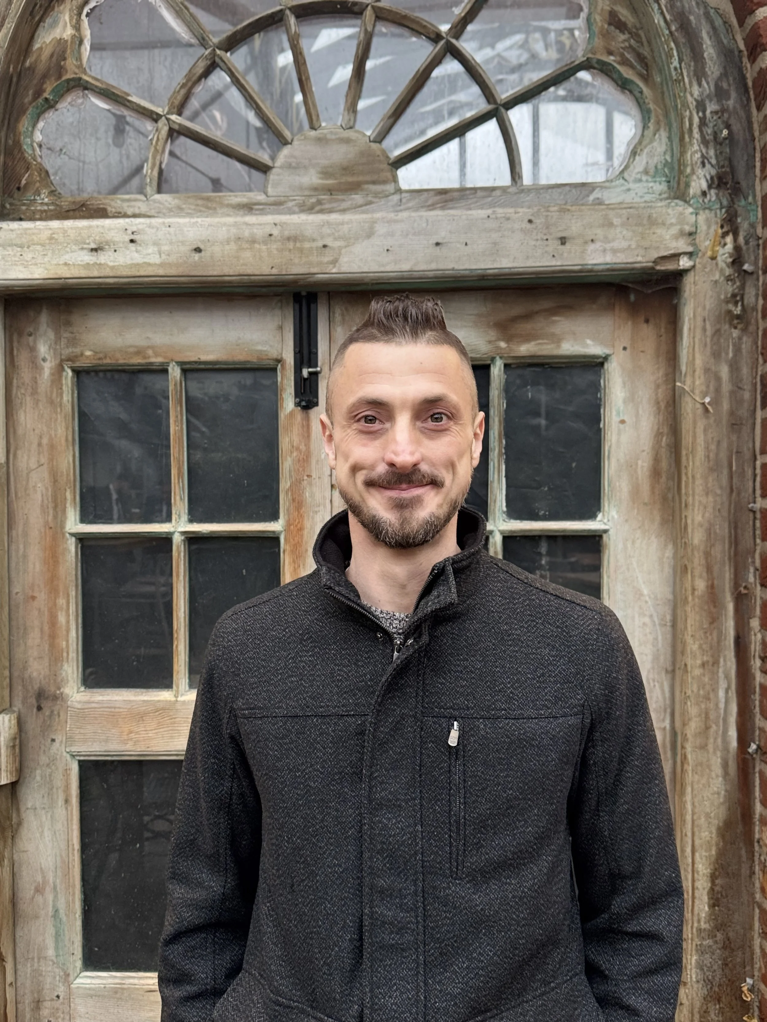 A young man sitting on a beige bench against a plain wall, wearing a gray polo shirt and cream-colored pants, with a neutral expression.