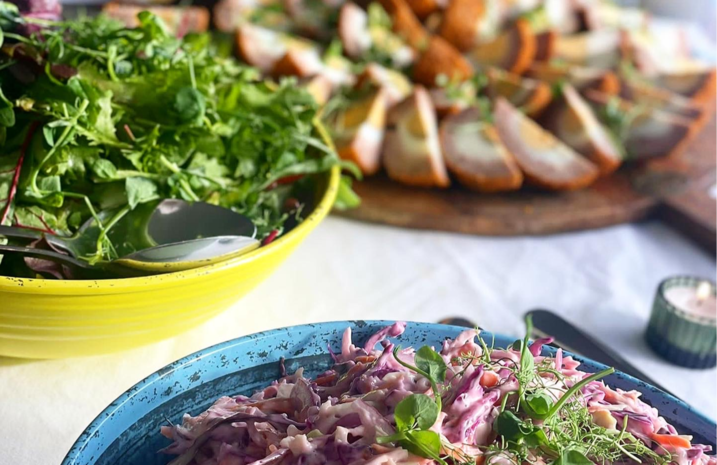 Colorful salad with leafy greens, shredded purple cabbage, and microgreens in a blue bowl, with other bowls of salad and sliced bread in the background.