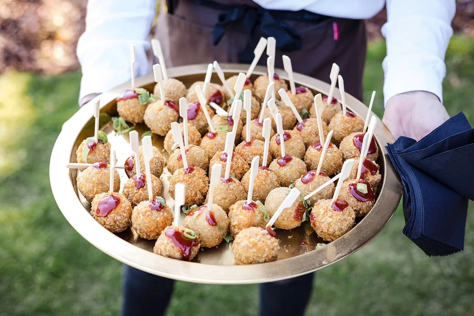 Person holding a tray of fried appetizers topped with sauce and garnishes, with a blue napkin in hand.