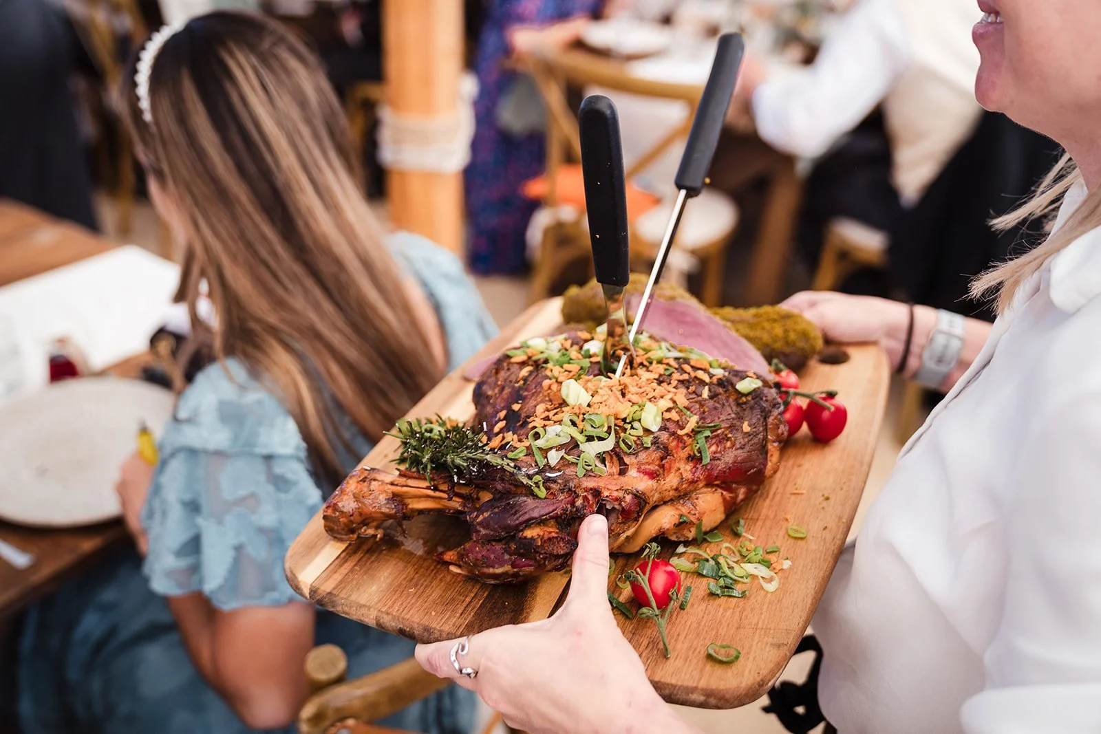 A person holding a wooden cutting board with a large roasted meat, topped with chopped green onions, nuts, and herbs, garnished with cherry tomatoes, in a restaurant setting.
