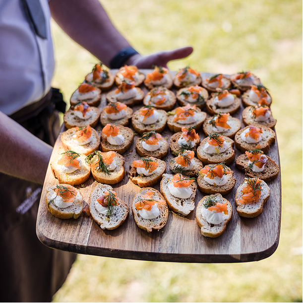 Person holding a large wooden tray with assorted canapés topped with smoked salmon, cream cheese, and herbs, outdoors on grass.