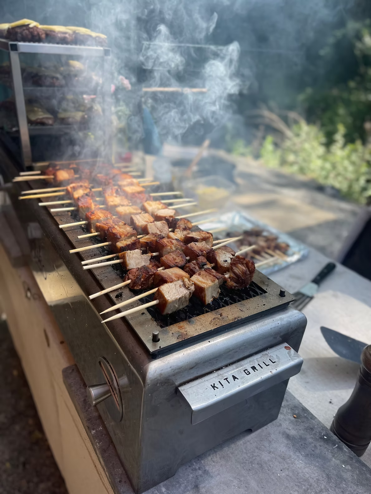Skewers of grilled meat cooking on a barbecue grill outdoors, with smoke rising from the heat.