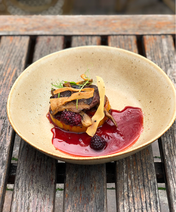 Gourmet dish with a piece of meat, berries, and sauce in a beige bowl on a wooden table.