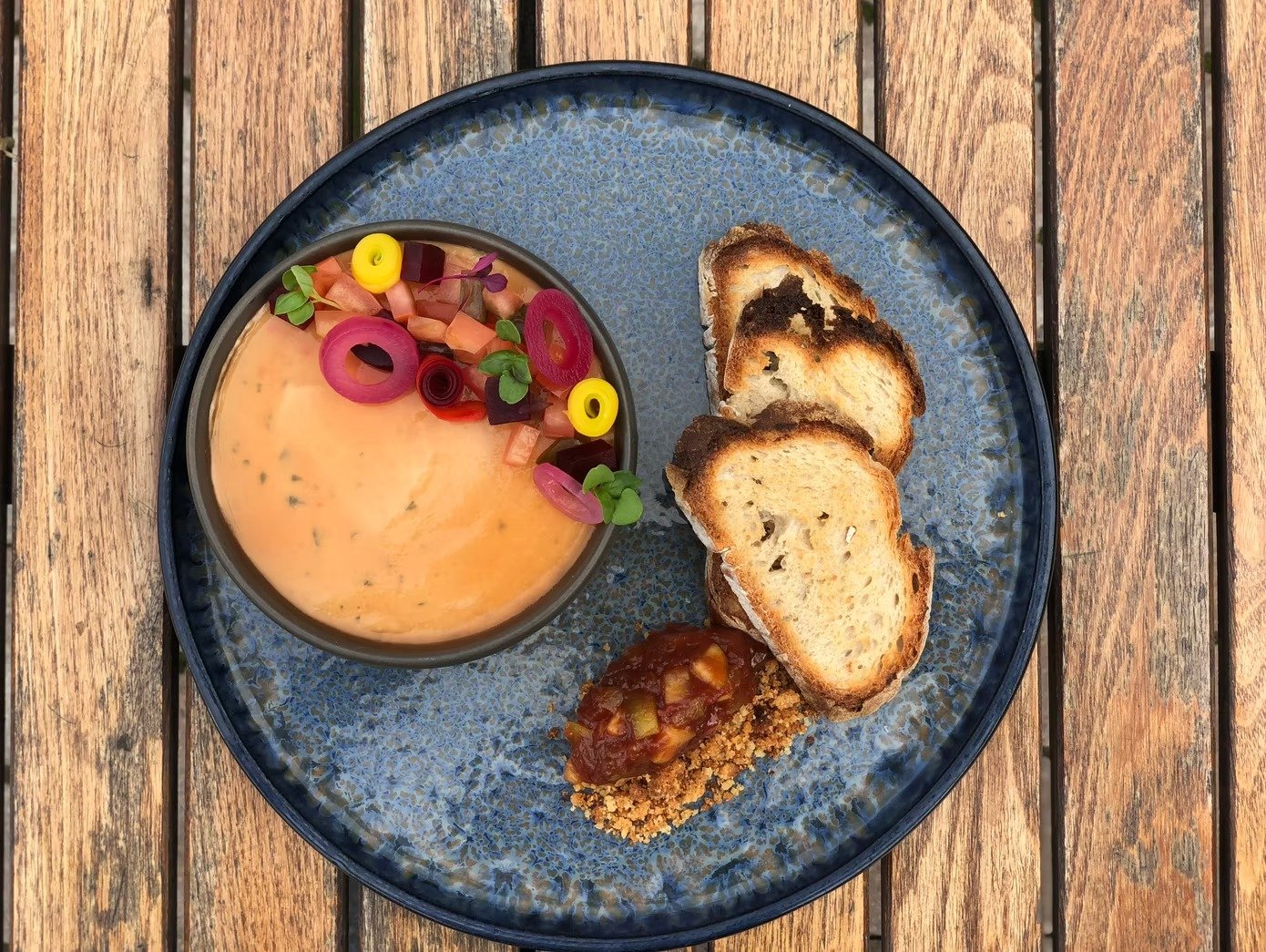 Plate with colorful dip topped with diced vegetables and pickled onions, toasted bread slices, and a spoonful of chili or stew, on a wooden table.