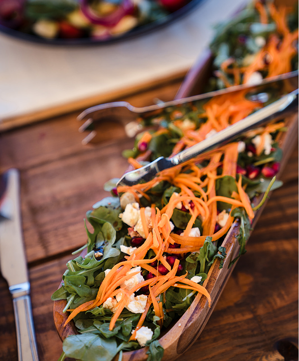 A fresh vegetable salad with greens, shredded carrots, feta cheese, and pomegranate seeds in a long wooden serving dish on a wooden table.