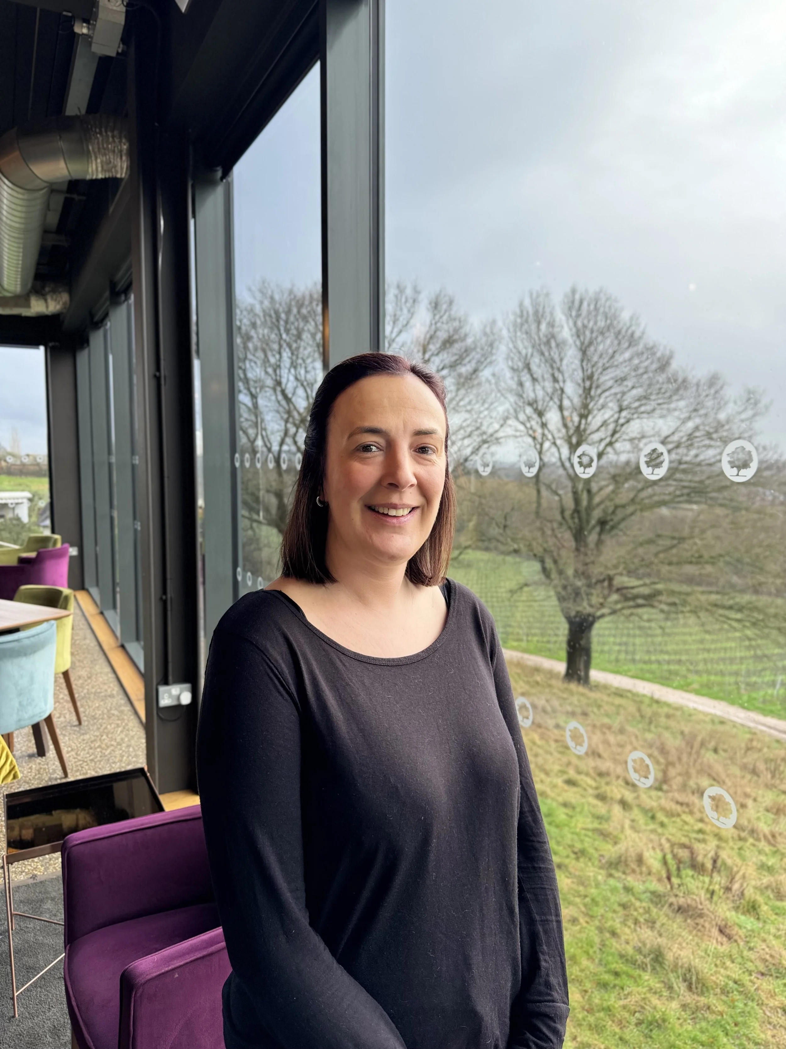A man with gray beard and short gray hair, dressed in a brown polo shirt and light-colored pants, sitting on a bench in a room with plain beige walls, looking thoughtfully to the side.