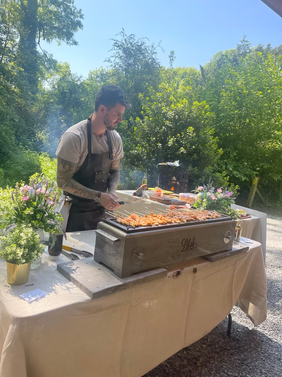 A man grilling skewers of food outdoors on a sunny day near a lush green forest, with a table decorated with flowers nearby.