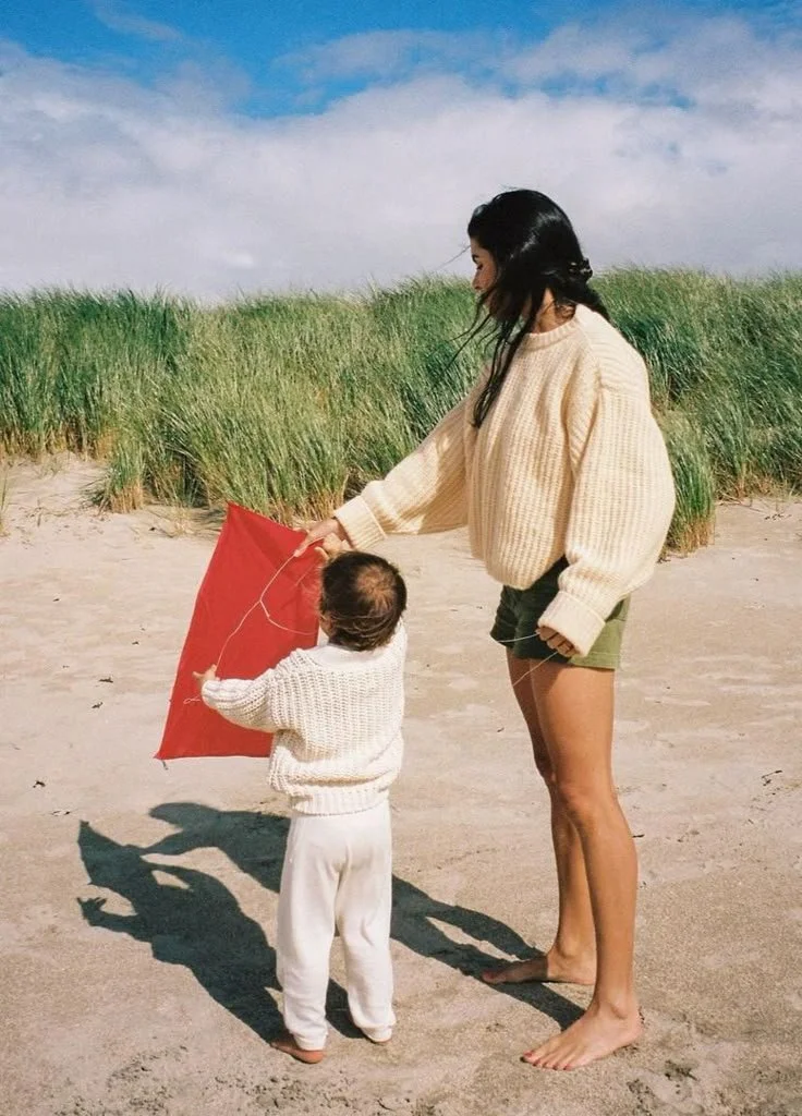 Une femme et un enfant qui tiennent un cerf-volant rouge sur la plage, avec des dunes et des herbes au fond, ciel nuageux en arrière-plan.
