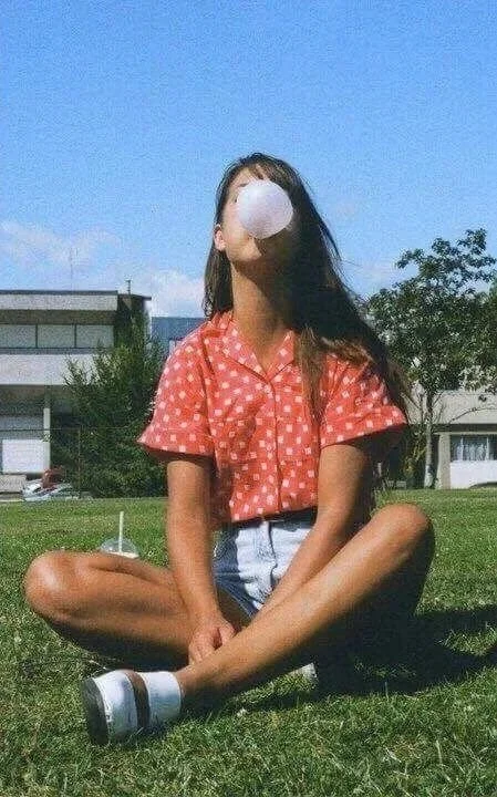 Jeune femme assise en plein air, portant un short et une chemise à pois rouges, soufflant une boule de savon.