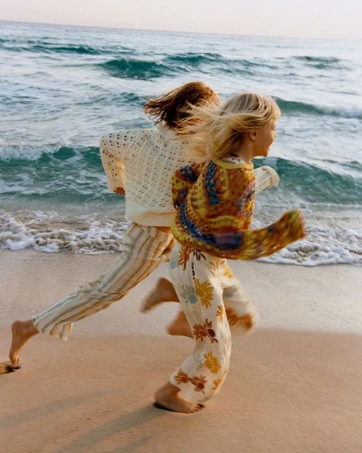 Deux enfants qui courent sur la plage près de la mer, avec des vagues en arrière-plan, vêtus de vêtements colorés et décontractés.
