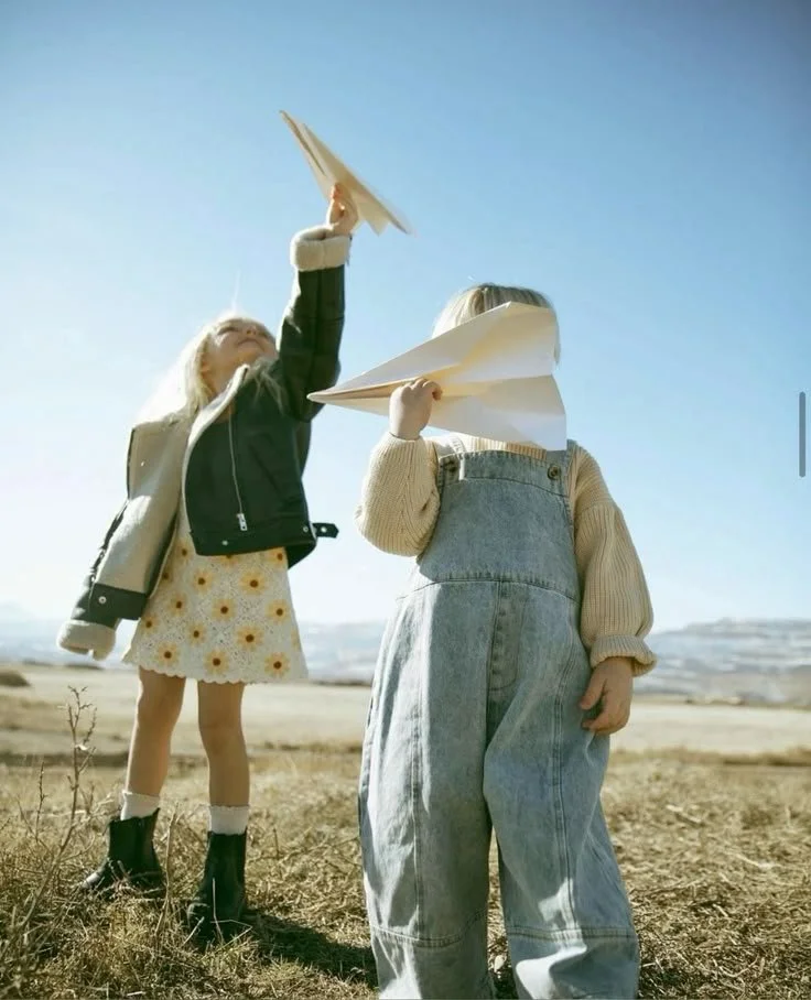 Deux enfants jouent avec des avions en papier dans un champ, avec un ciel clair en arrière-plan.