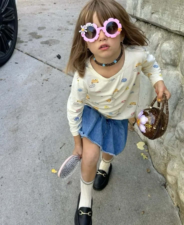 Une jeune fille avec des lunettes de soleil roses en forme de fleurs, portant un sweat blanc avec des motifs, une jupe en denim, des chaussettes beiges et des mocassins noirs, tenant une petite trousse et un sac à main en forme de lanterne ornée de fleurs, debout devant un mur en pierre sur le trottoir.