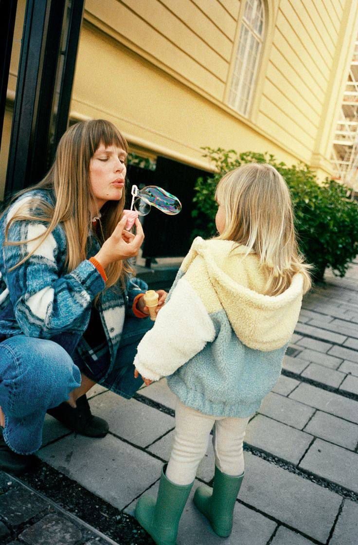 Une femme Toujours Plus, Jamais Assez souffle sur une bulle de savon que tient une fillette, en extérieur devant une maison en bois.