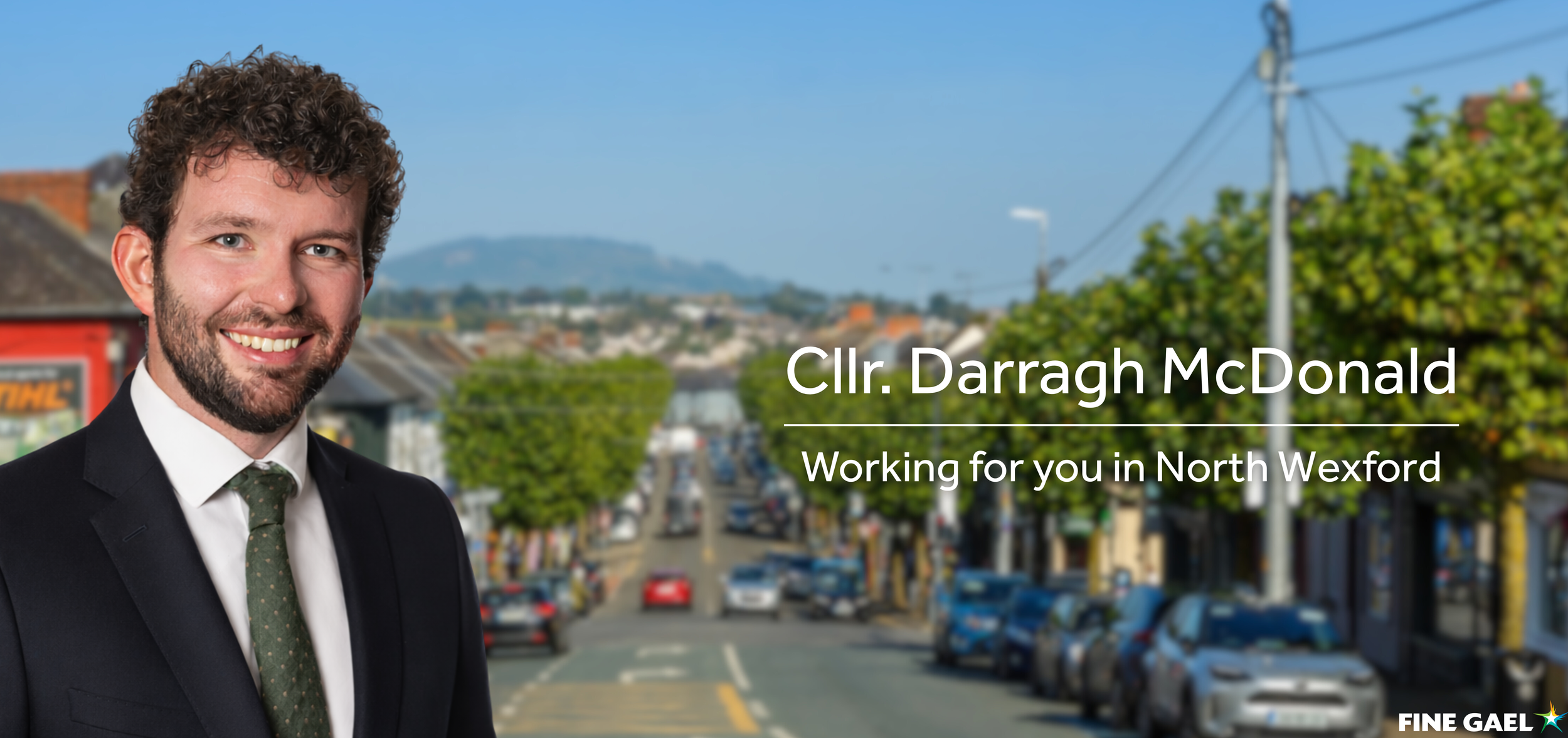 Portrait of Councilor Darragh McDonald wearing a suit and tie, with a busy street in North Wexford in the background, showing parked cars, trees, and buildings under a clear sky.