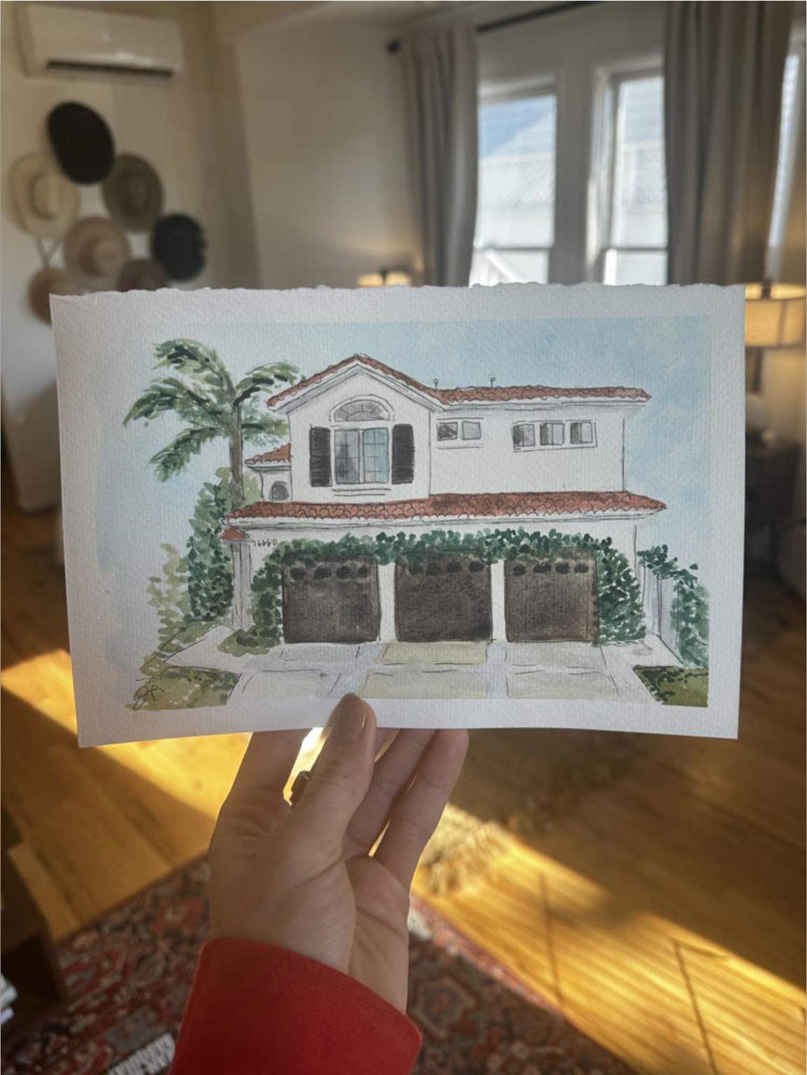 Hand-painted watercolor of a two-story house with three garage doors, a red tile roof, and surrounding greenery, held up indoors with a living room in the background.