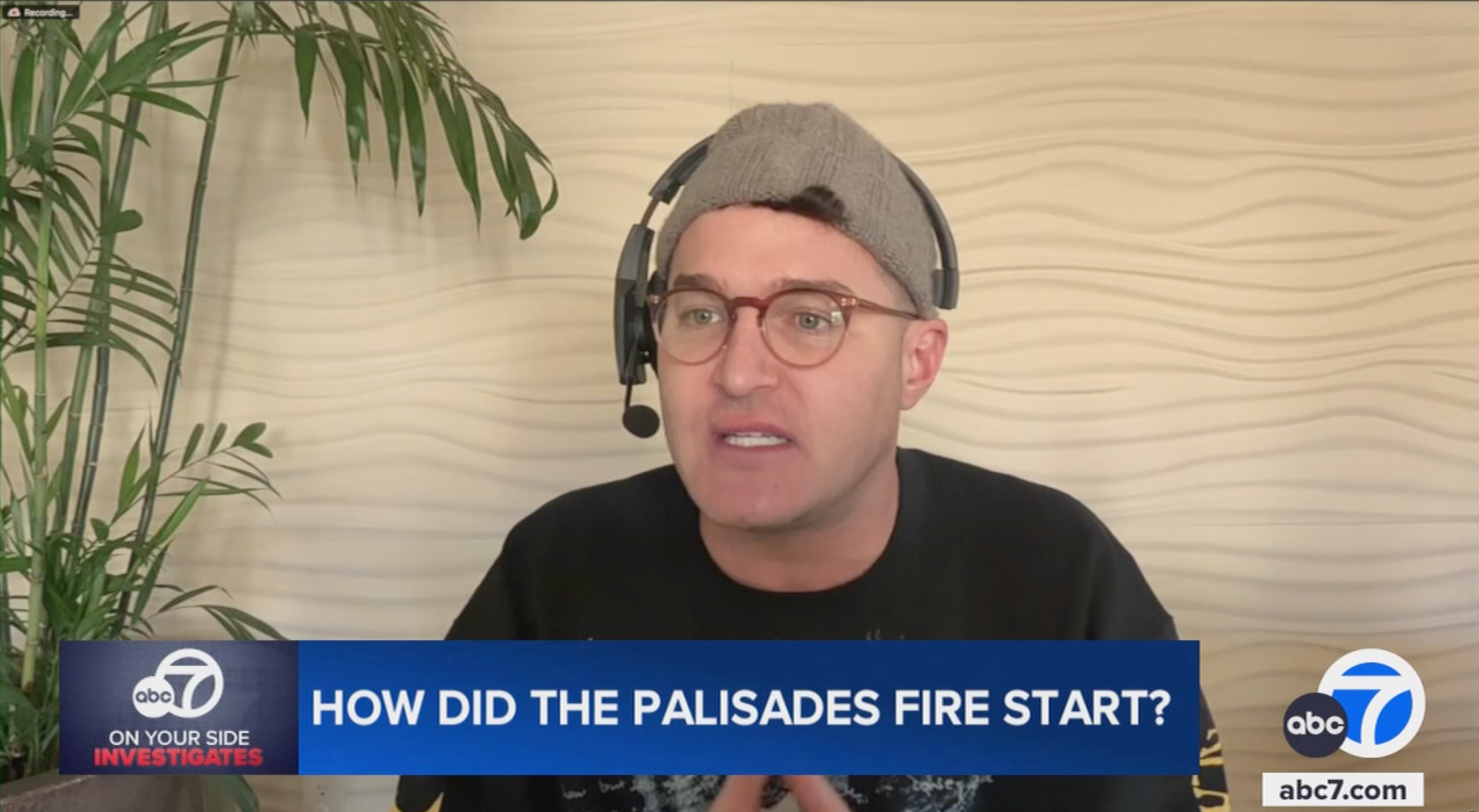 A man wearing glasses, a gray beanie, and a headset with a microphone is speaking on a news broadcast. There is a potted plant on the left and a light-colored wavy textured wall behind him. The news banner at the bottom reads "How did the Palisades fire start?" from ABC 7 News.