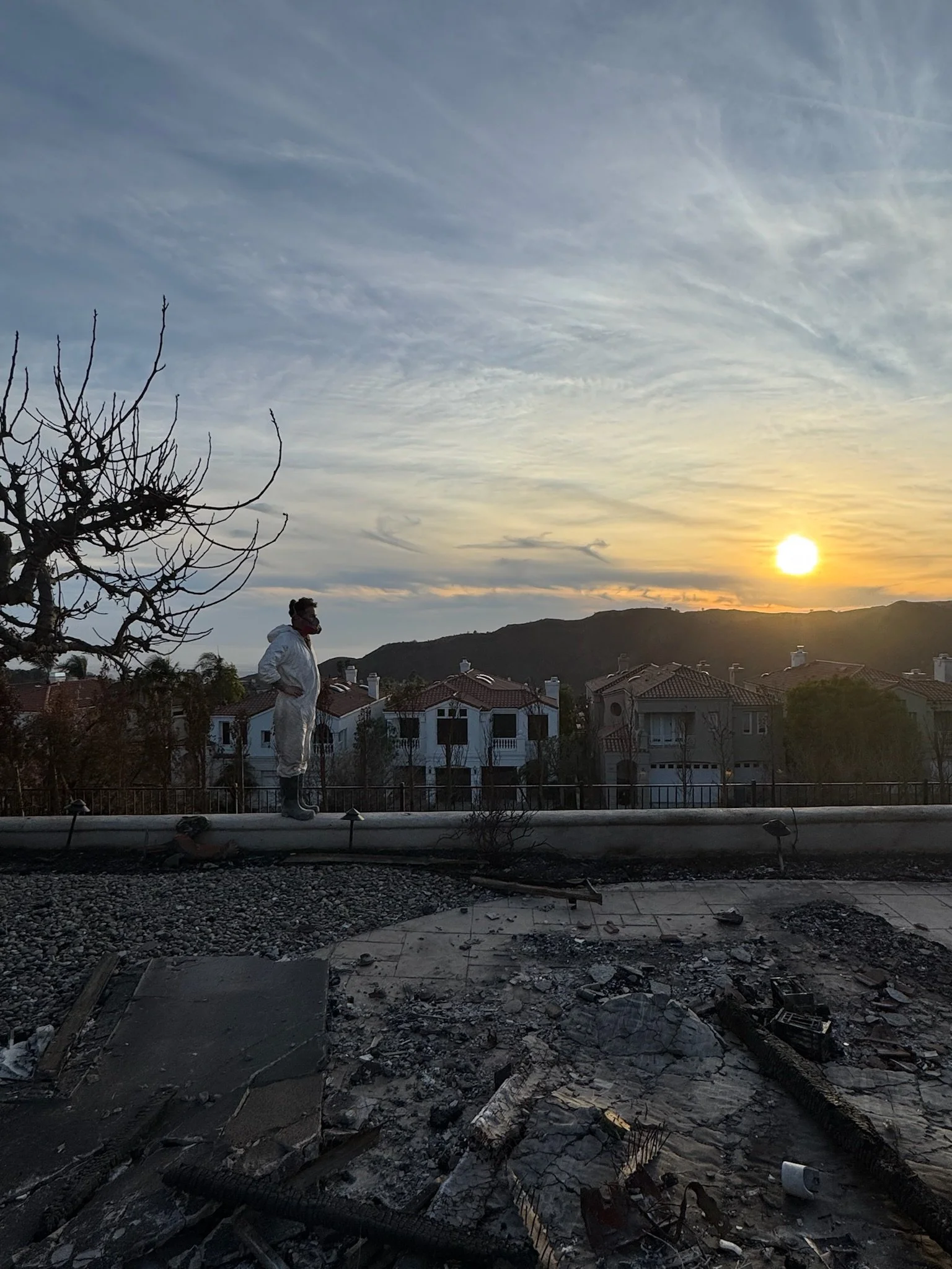 A person dressed in white protective suit stands amid the charred remains of a burned roof at sunset, with houses and hills in the background.