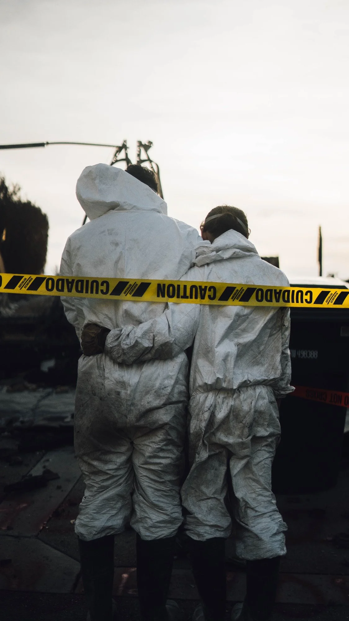 Two individuals in white hazmat suits stand together behind yellow caution tape labeled "CUIDADO" and "CAUTION" at what appears to be a fire scene.