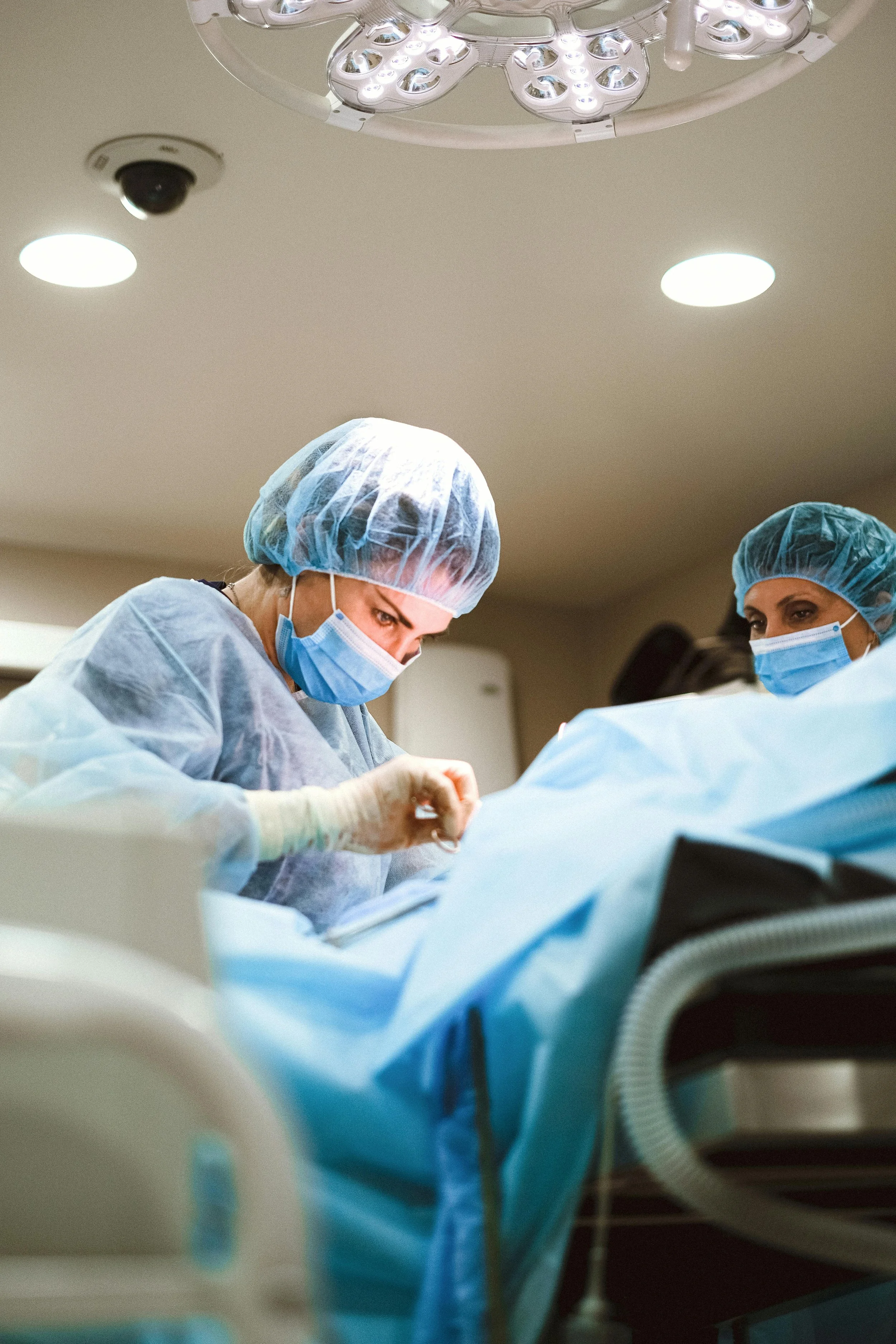 Two surgeons in scrubs and masks performing surgery in an operating room, under bright surgical lights.