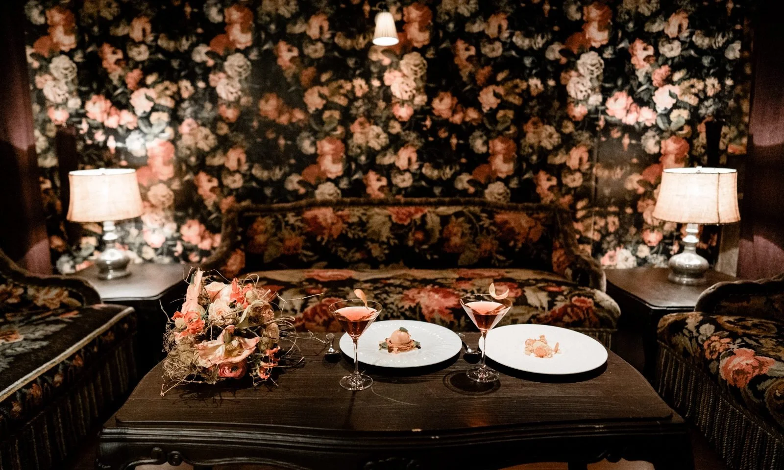 A cozy, vintage-style living room features floral wallpaper, matching floral upholstery on sofas, two side tables with lamps, and a dark wooden coffee table with flowers, two cocktails, and desserts.