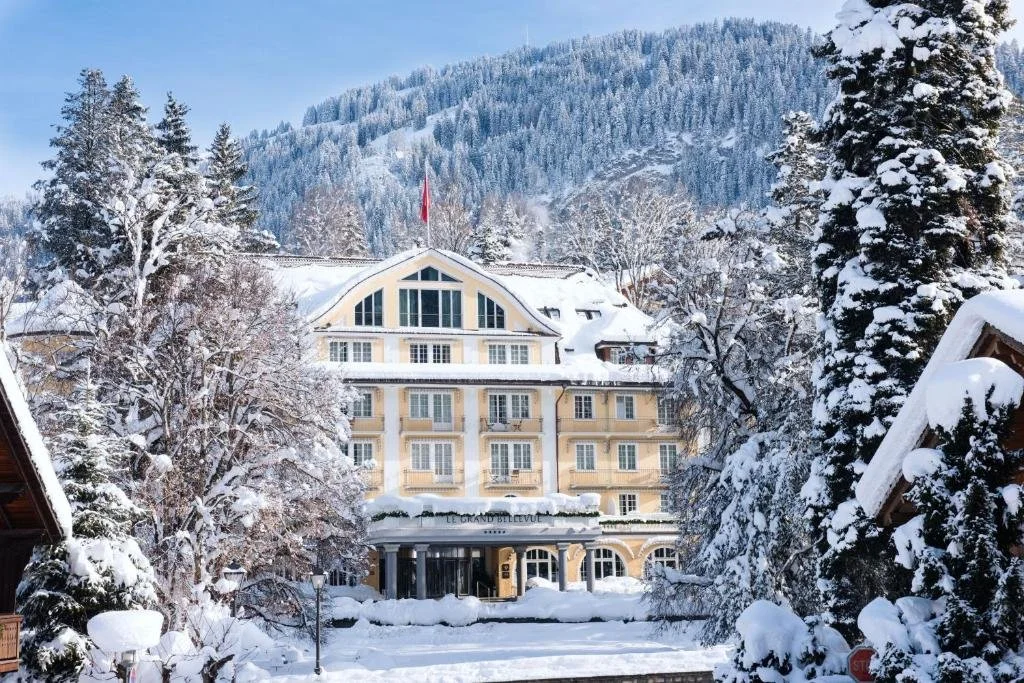 A large hotel surrounded by snow-covered trees and mountains, with a cloudy sky in the background.