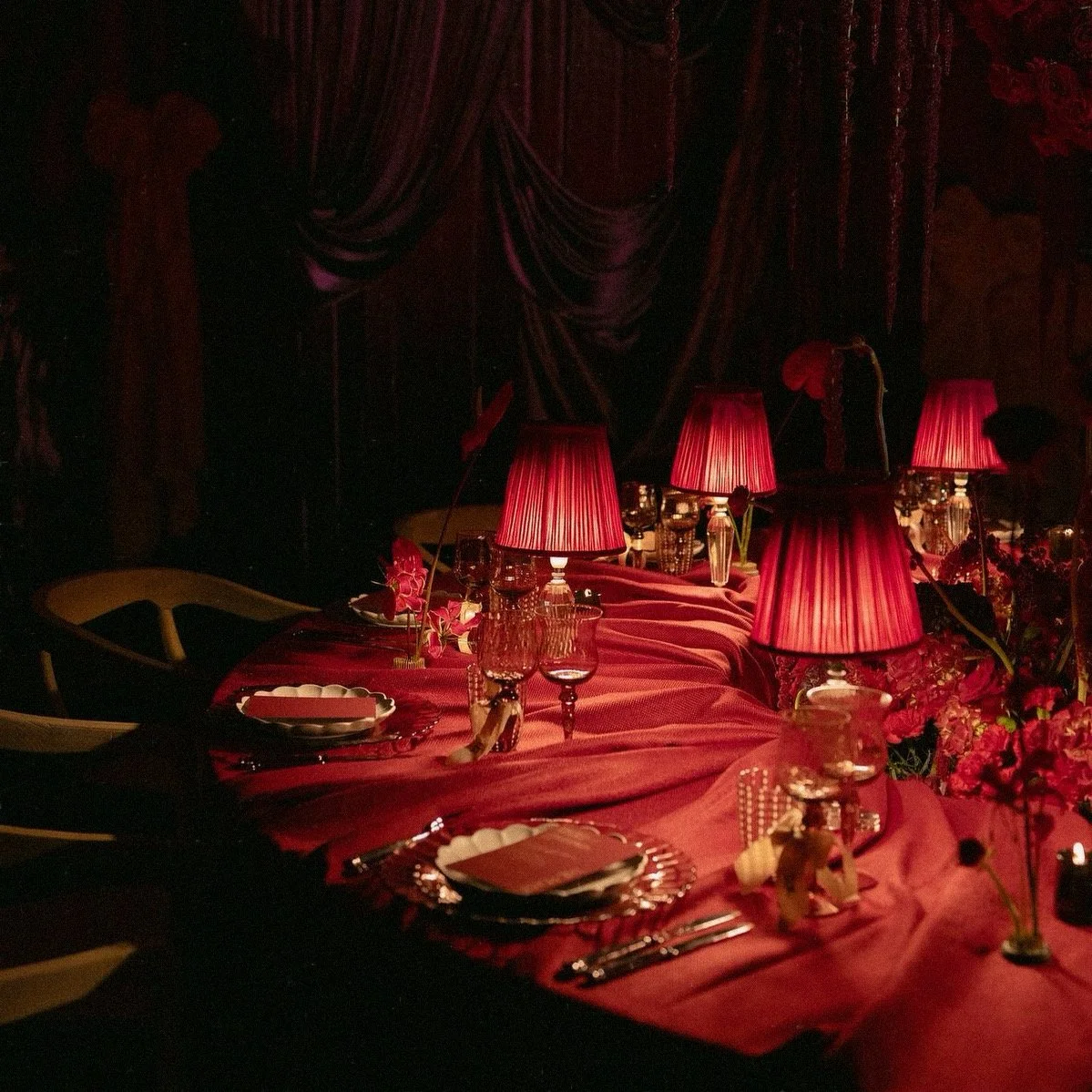 A dimly lit dining table decorated with pink tablecloths, pink lampshades, glasses, plates, and floral centerpieces, suggesting an elegant, romantic setting.