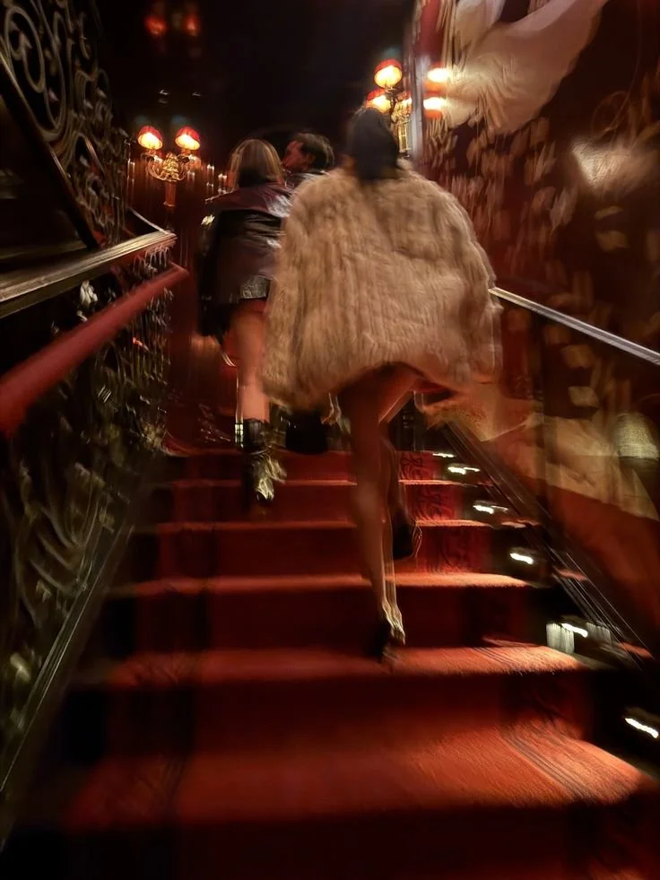 People walking up a dark, ornate staircase with red carpet and red wall lamps.