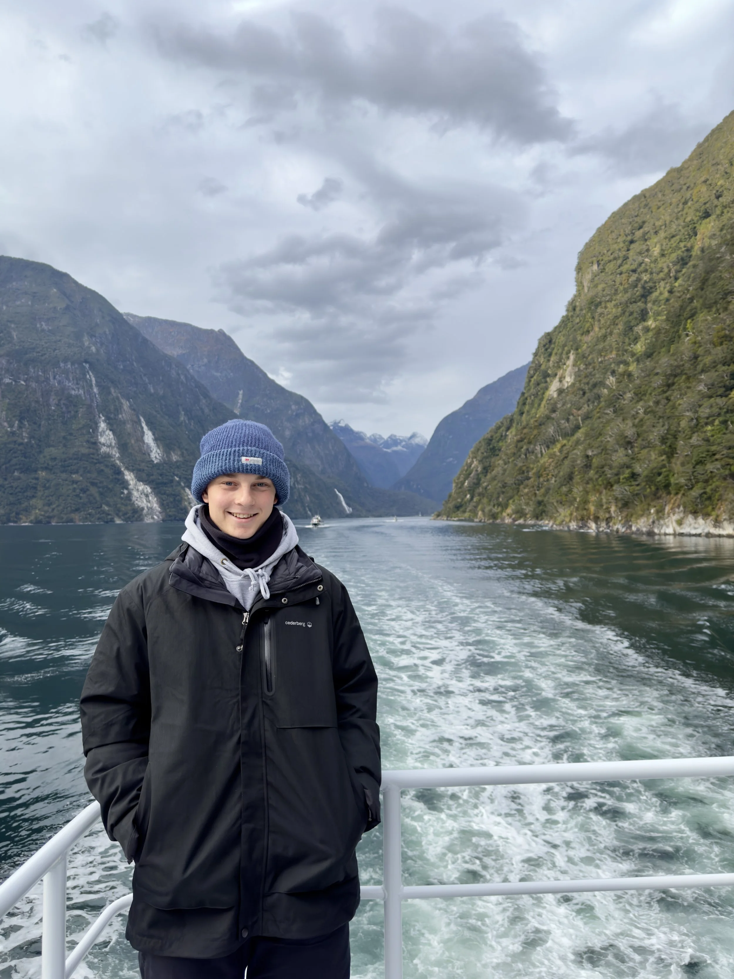 A smiling young man in a black jacket and blue beanie standing on the deck of a boat on a fjord, with mountains and waterfalls in the background under a cloudy sky.