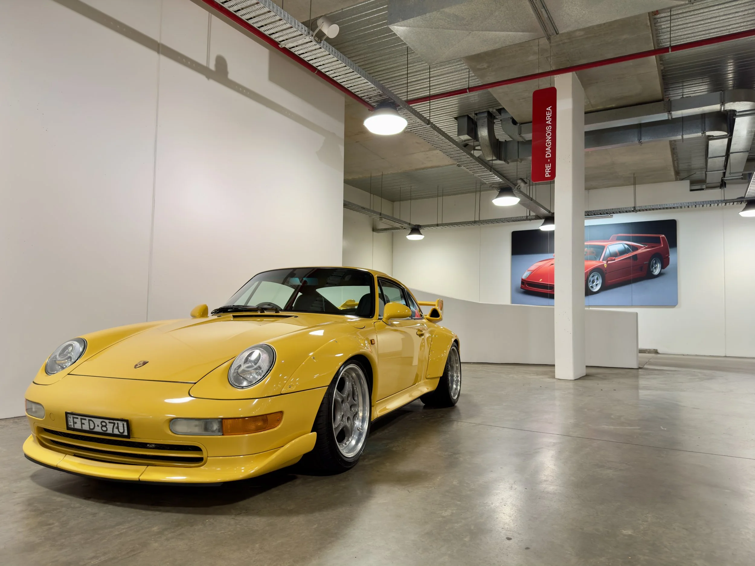 A yellow Porsche 911 race car displayed in an indoor showroom with a photo of a red sports car on the wall behind it.