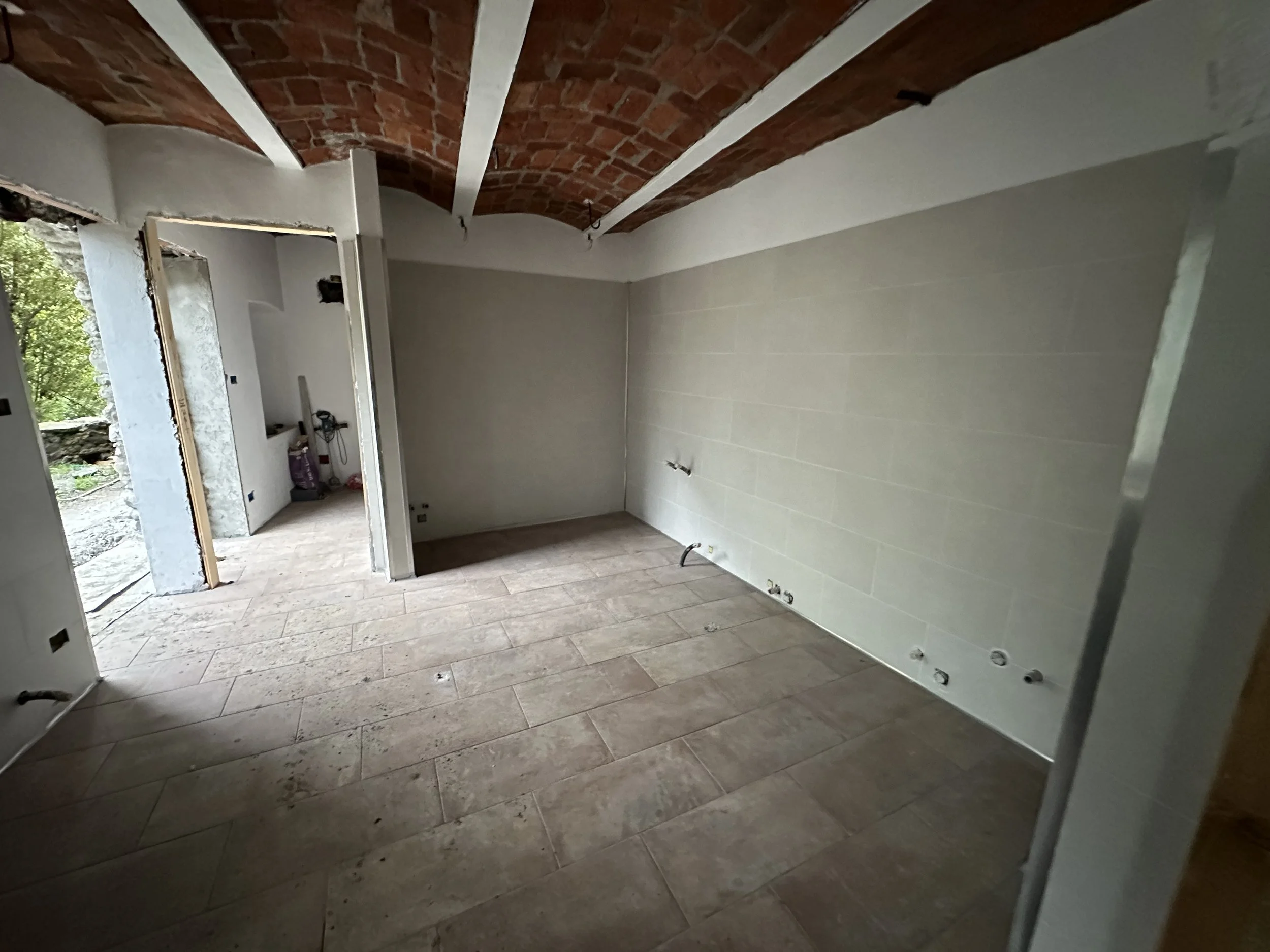 Stanza in fase di ristrutturazione con pareti chiare, soffitto con travi di legno e pavimento in piastrelle di marmo. Una porta aperta su una stanza adiacente, con vista su un'area esterna con alberi.