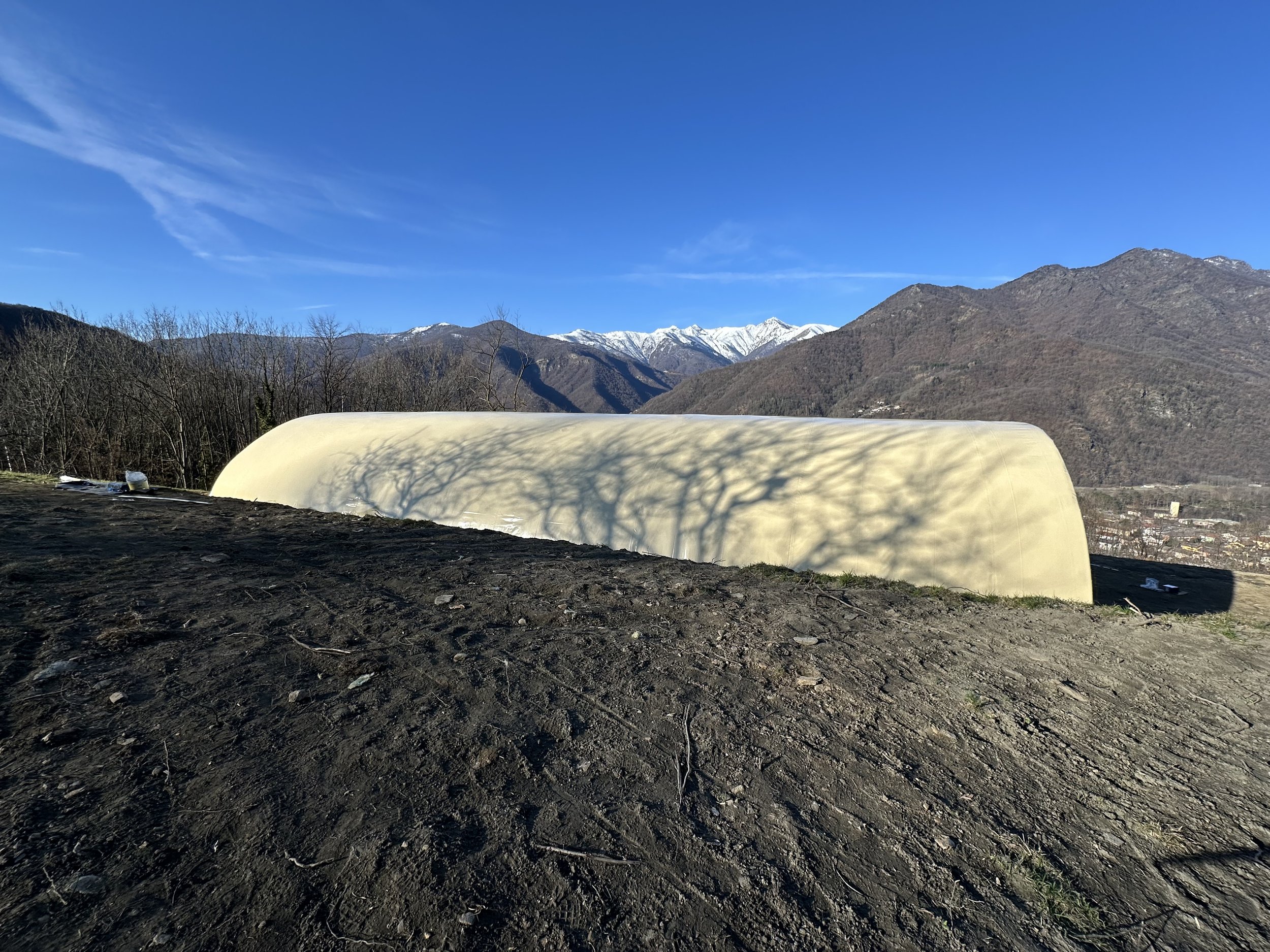 Struttura di colore giallo chiaro con ombra di un albero sulla sua superficie, situata in una regione montuosa con montagne innevate sullo sfondo, cielo blu e terreno terrazzato.