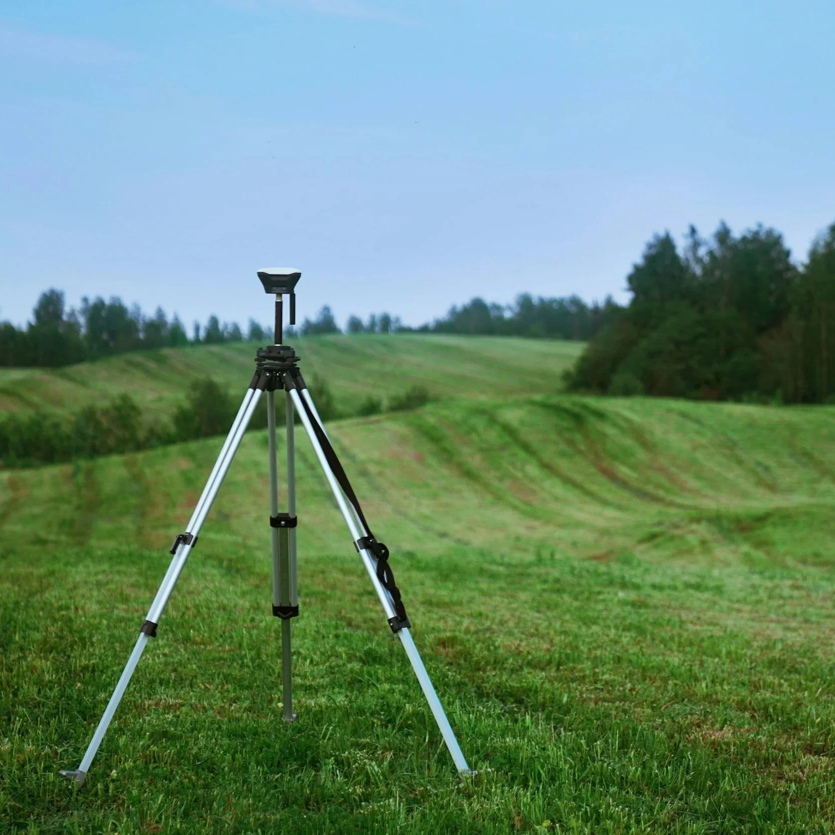Trepied su un prato verde con un paesaggio collinare e alberi sullo sfondo, sotto un cielo azzurro.