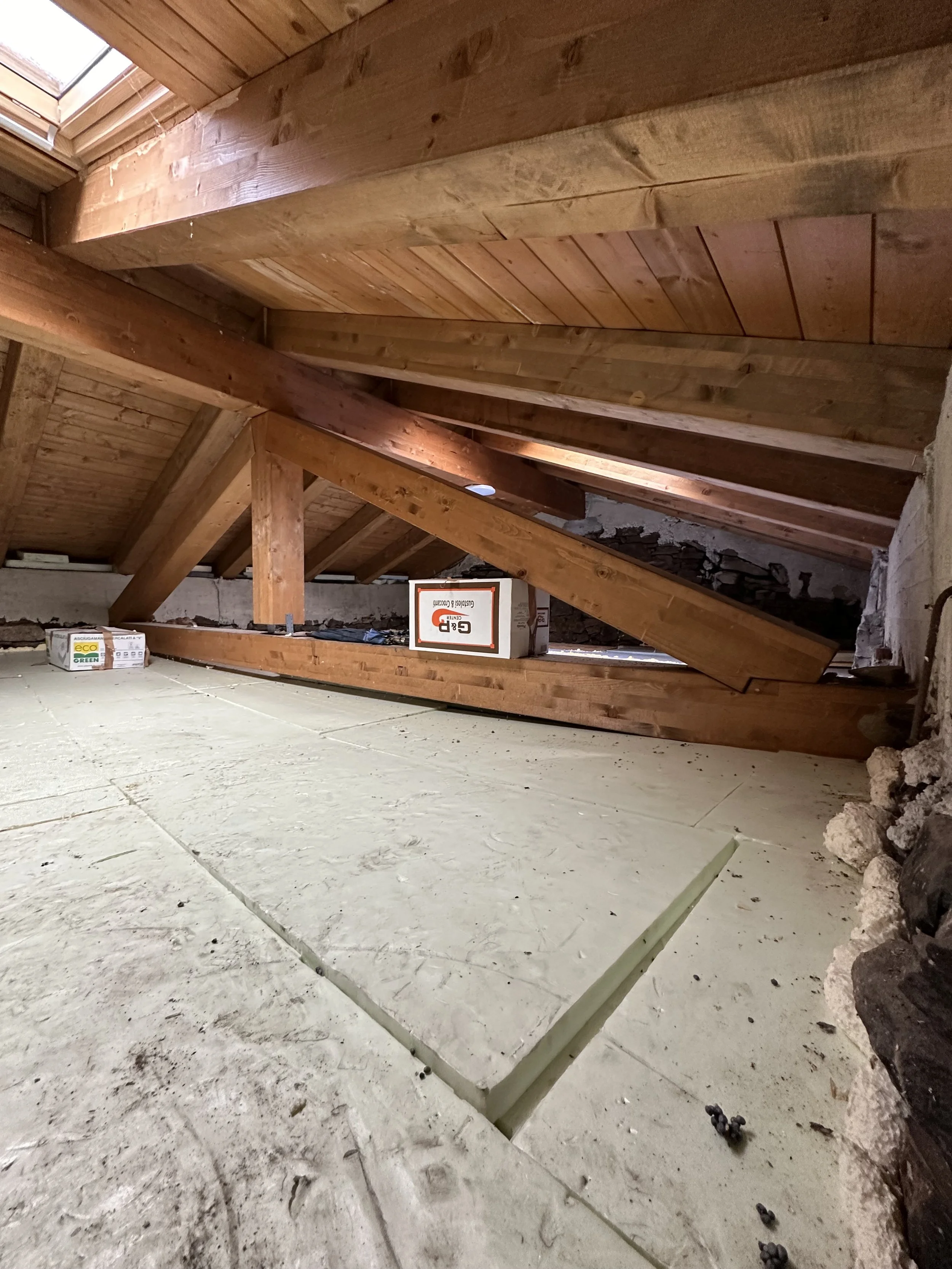 Interno di un sottotetto con travi di legno e pavimento in calcestruzzo, con alcune scatole di materiali da costruzione e una grande crepa nel pavimento.