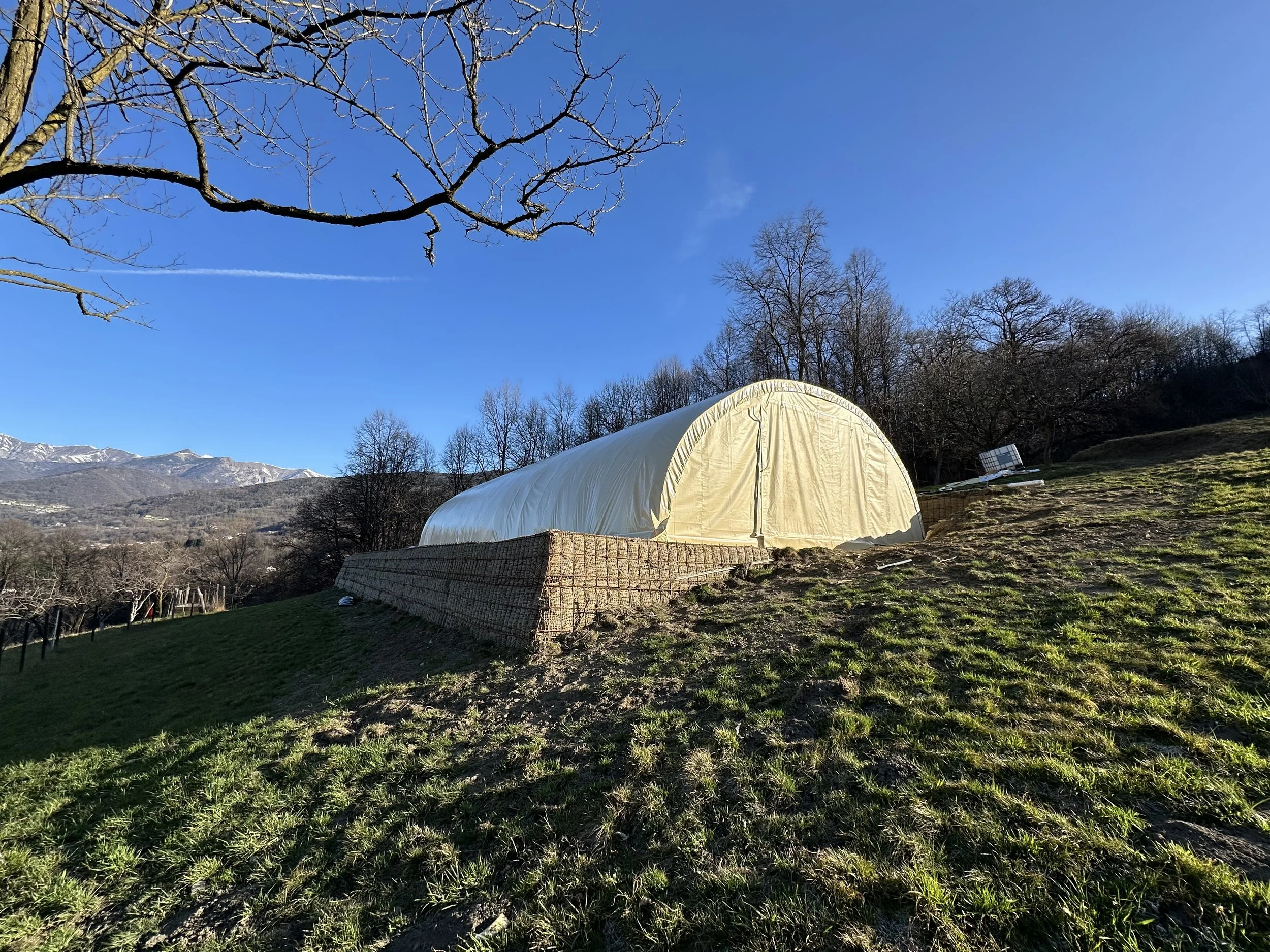 Tenda bianca in un paesaggio collinare con alberi spogli e montagne sullo sfondo sotto un cielo blu.