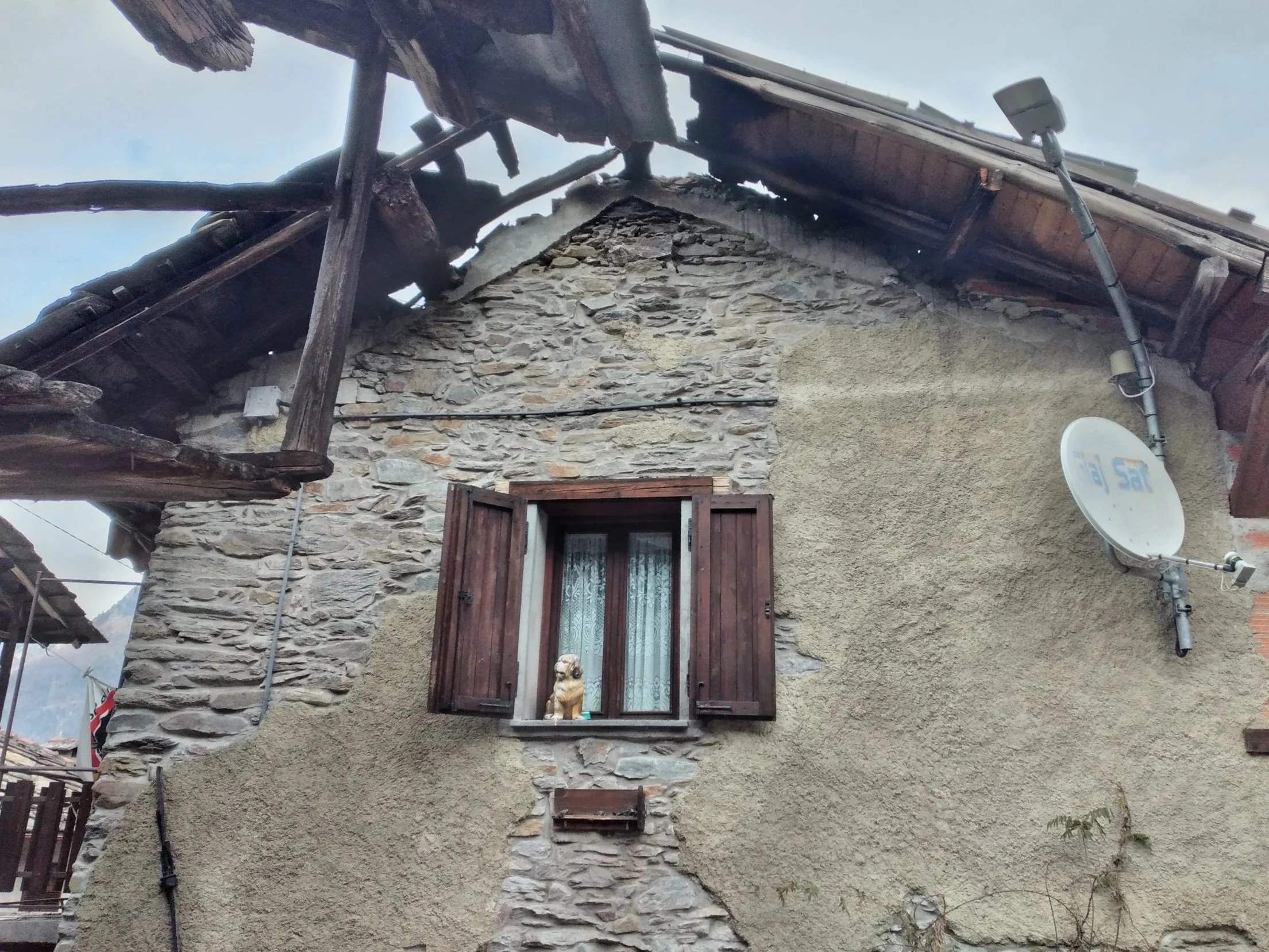 Facciata di una casa in pietra con finestra con persiane di legno, satellite e tetto danneggiato.