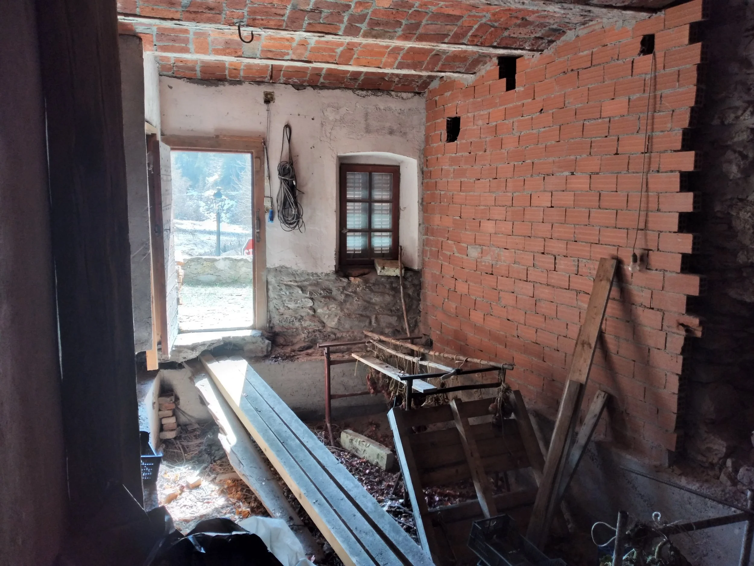 Interno di una stanza in costruzione con mattoni rossi e pareti in cemento, finestra con vista sul paesaggio esterno innevato, attrezzi e materiali da costruzione sparsi sul pavimento.