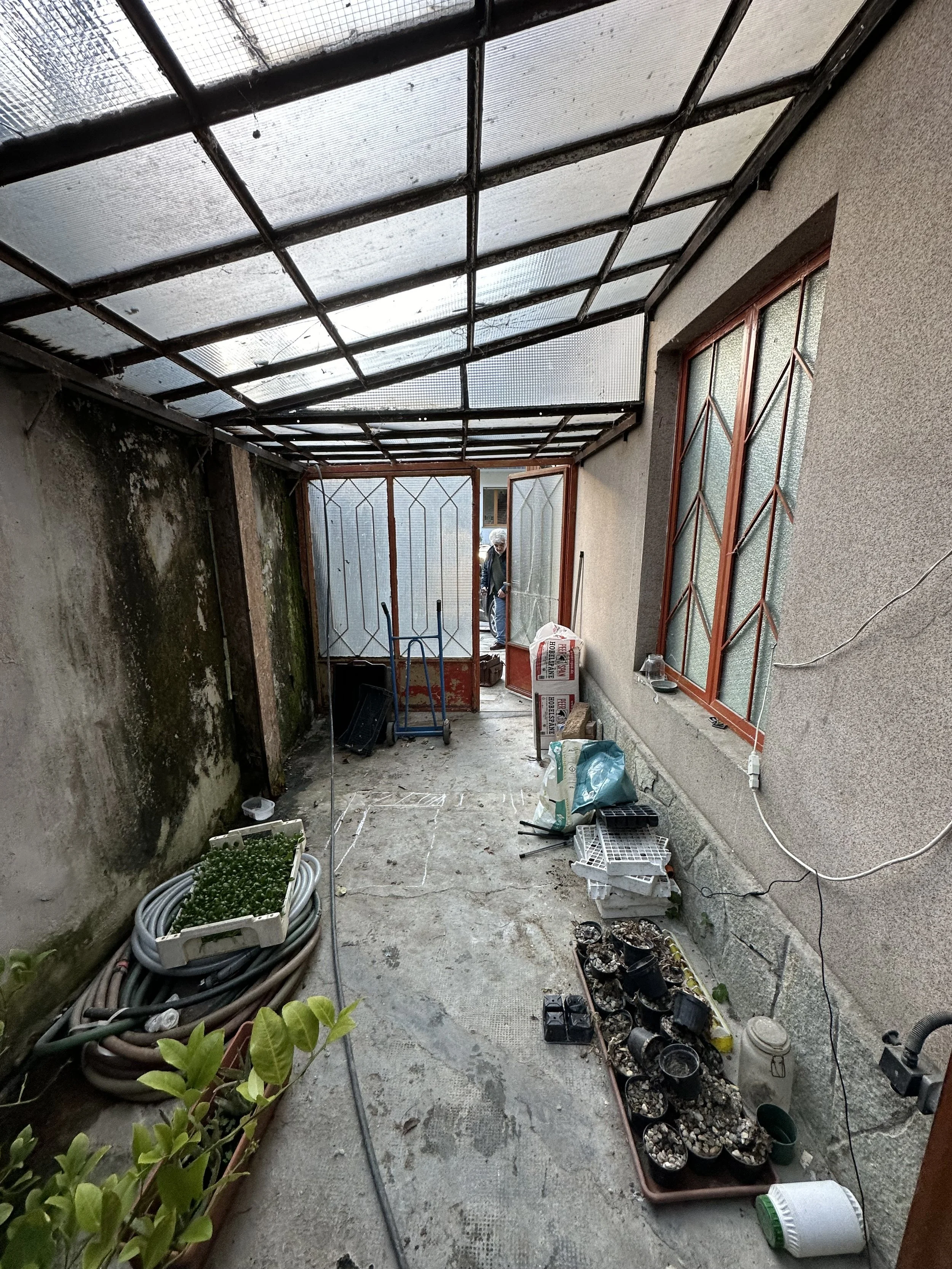 Veranda coperta con strutture di metallo e plastica trasparente, muri con muffa, piante in vaso, attrezzi da giardinaggio e scatole di materiali in fai-da-te.